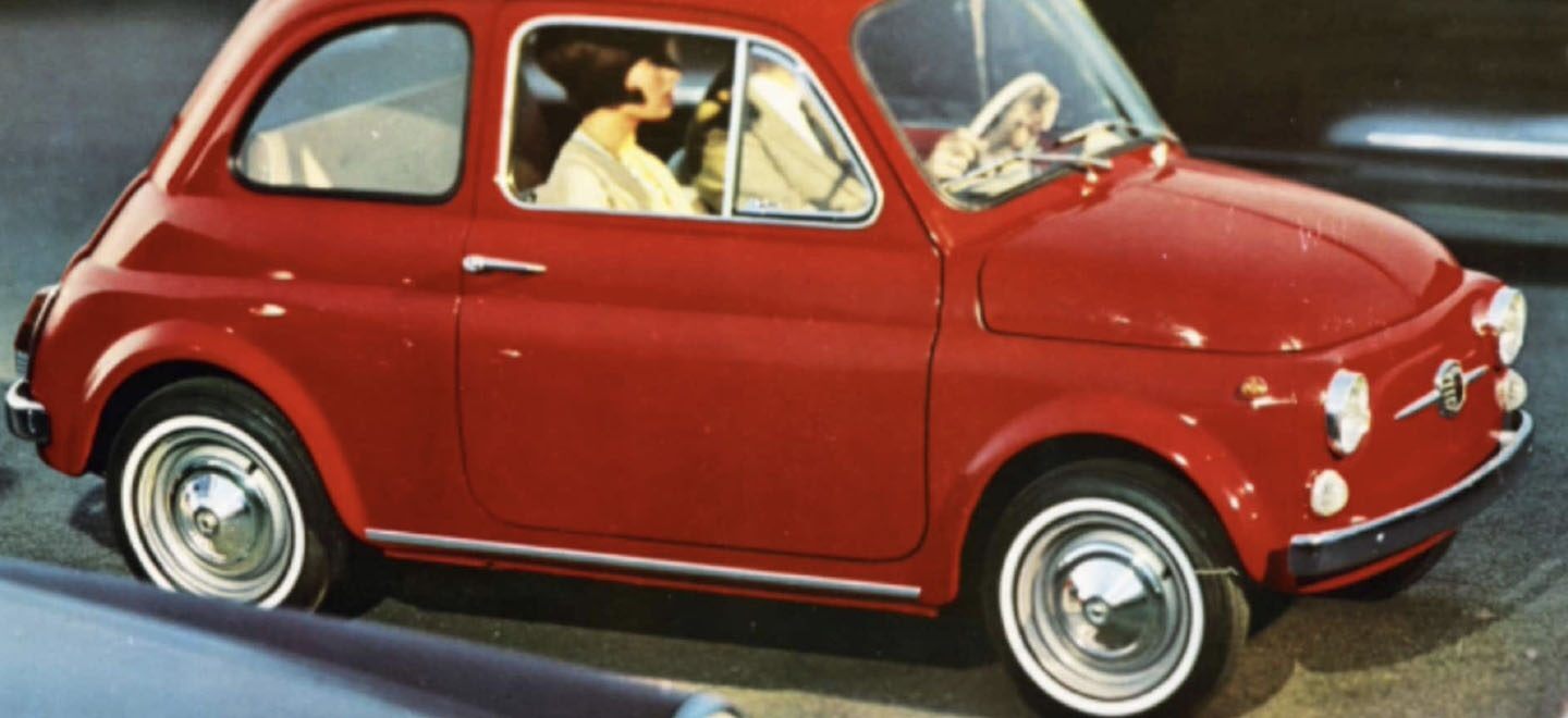Se muestra una foto vintage de un Fiat 500 clásico circulando por la calle con una pareja delante.