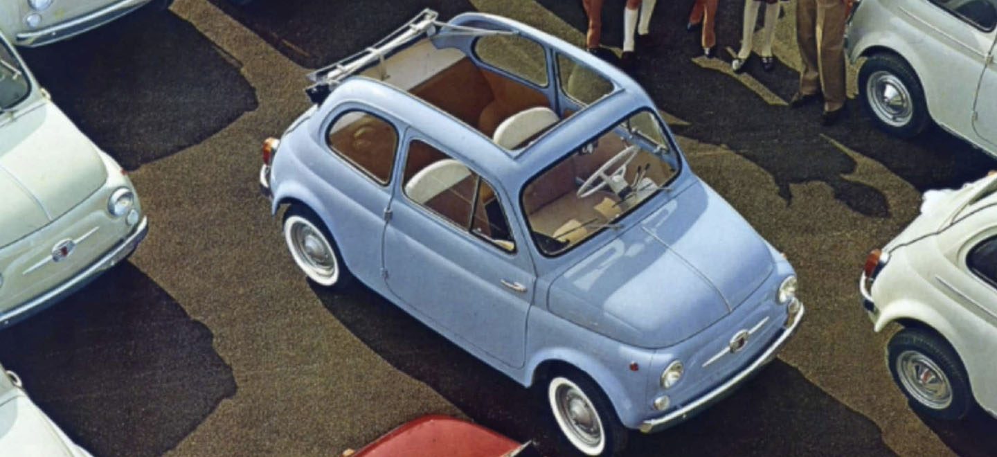 Se muestra un dibujo "vintage" desde arriba de un Fiat Cabrio clásico azul cielo estacionado entre varios otros vehículos Fiat clásicos.