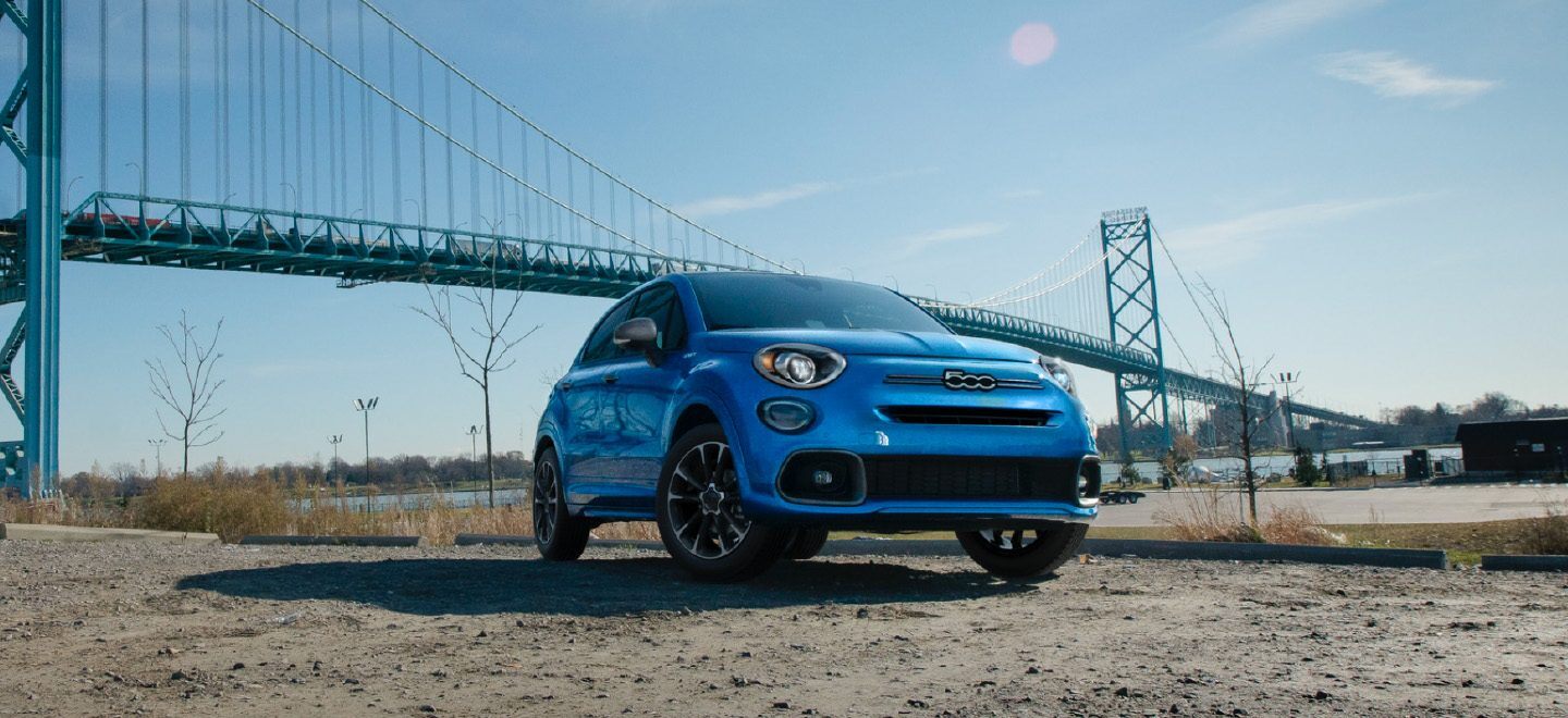 Display A blue 2022 Fiat 500X Yacht Club Capri parked in a clearing with a suspension bridge in the background.