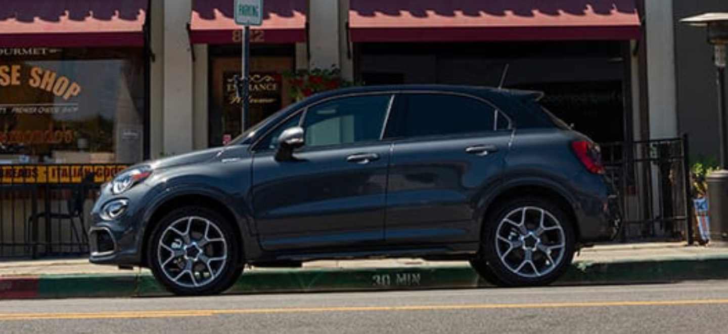 Se muestra una vista de perfil de un Fiat 500X Sport 2021 estacionado delante de una pintoresca tienda de quesos.